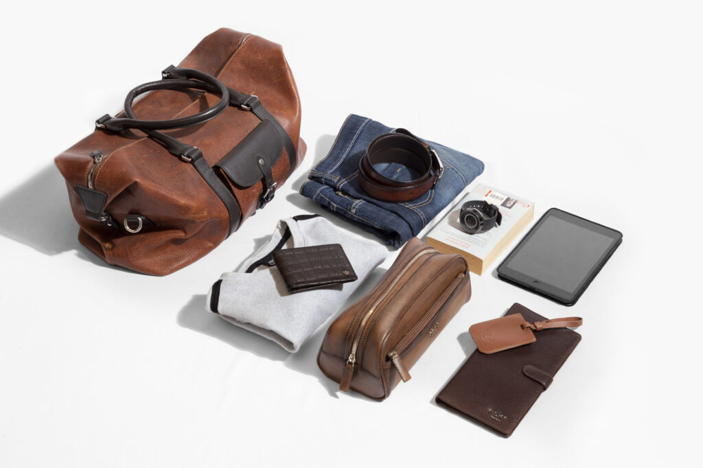 A flatlay photo of Men's Travel Duffle Bag in brown with tshirts, jeans, wallet , belt, watch, passport cover and ipad